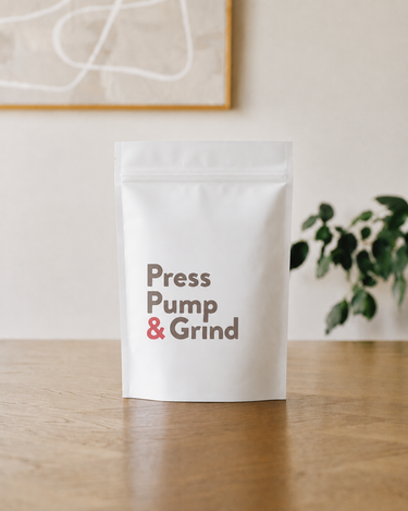 White packaging with 'Press Pump & Grind' text on a wooden surface with a plant in the background