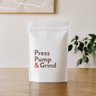 White packaging with 'Press Pump & Grind' text on a wooden surface with a plant in the background